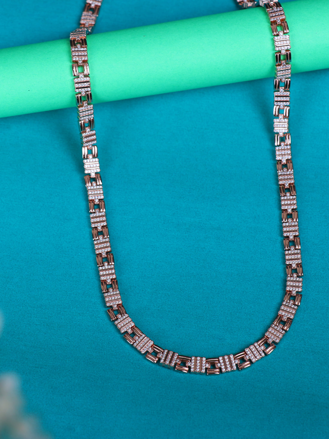 A silver chain necklace with rectangular links set with small clear stones, draped over a mint green cylindrical prop on a bright turquoise background.