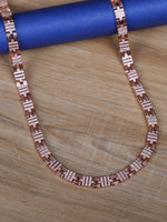 A rose gold color silver chain necklace with rectangular links set with small clear stones, displayed on a light wood surface next to a blue cylindrical prop.