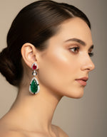 Woman wearing long silver earrings with red and green stone drops and sparkling halo accents.